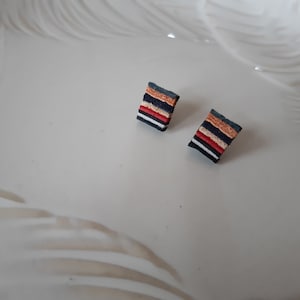 Könnte beinhalten: Ein Paar kleine quadratische Ohrringe mit einem geschichteten Design. Die Ohrringe sind aus dunkelblauem, schwarzem, weißem, rotem und braunem Material.