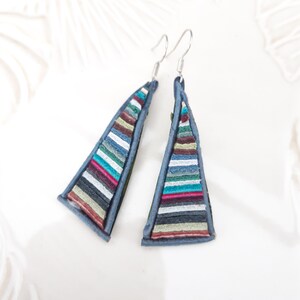 May include: A pair of blue triangle-shaped earrings with a multi-colored striped design. The stripes are made of different shades of blue, green, red, white, and brown.