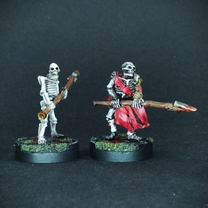 May include: Two painted skeleton figures holding weapons. The skeleton on the left is holding a wooden staff. The skeleton on the right is holding a spear with a red and brown handle. Both skeletons are standing on black bases with green and brown accents.