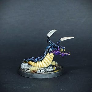 Pode incluir: Uma miniatura pintada à mão de uma criatura vermiforme roxa e azul com partes do corpo segmentadas amarelas. A criatura tem duas asas brancas e está em uma base preta com pedras marrons e cinzas.