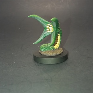 May include: A green and yellow painted miniature of a creature with a long, segmented tail and a large, open mouth with rows of teeth. The creature is standing on a small, round base.