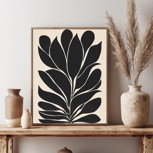 Op de afbeelding: Zwart-witte abstracte kunstprint van een gestileerde plant met grote bladeren. De print is ingelijst en wordt tentoongesteld op een houten plank met twee vazen.