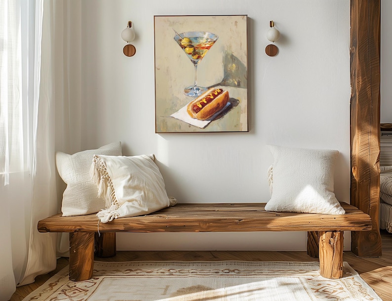 May include: A painting of a martini and a hot dog on a white napkin. The painting hangs above a wooden bench with two white pillows. The bench is made of rough-hewn wood and has a natural finish. Two wall sconces are mounted on the wall.