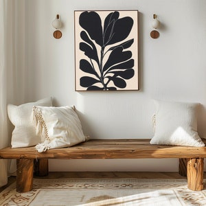 Op de afbeelding: Een houten bank met twee witte kussens staat voor een witte muur met een zwart-witte abstracte bloemenprint die eraan hangt.