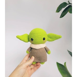 May include: A green and brown crocheted amigurumi toy resembling a popular fictional character. The toy has large ears, a small mouth, and big eyes. The toy is being held in a hand.