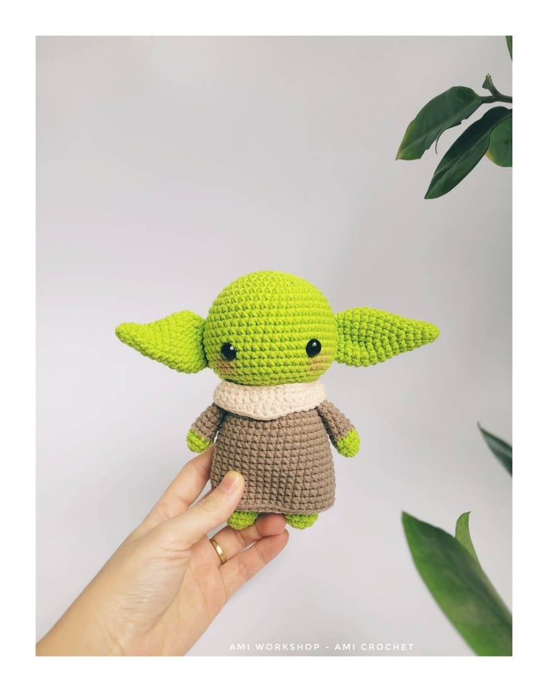 May include: A green and brown crocheted amigurumi toy resembling a popular fictional character. The toy has large ears, a small mouth, and big eyes. The toy is being held in a hand.