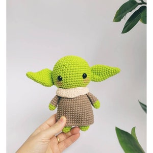 May include: A green and brown crocheted plush toy resembling a popular fictional character. The toy has large ears, a small mouth, and big eyes.