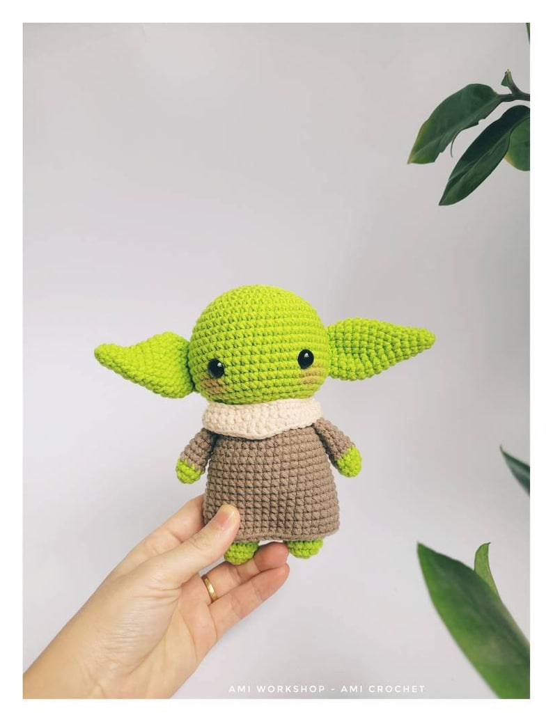 May include: A green and brown crocheted plush toy resembling a popular fictional character. The toy has large ears, a small mouth, and big eyes.