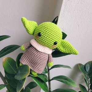 May include: A green crocheted amigurumi toy with brown clothing and large black eyes. The toy is sitting on a green plant.
