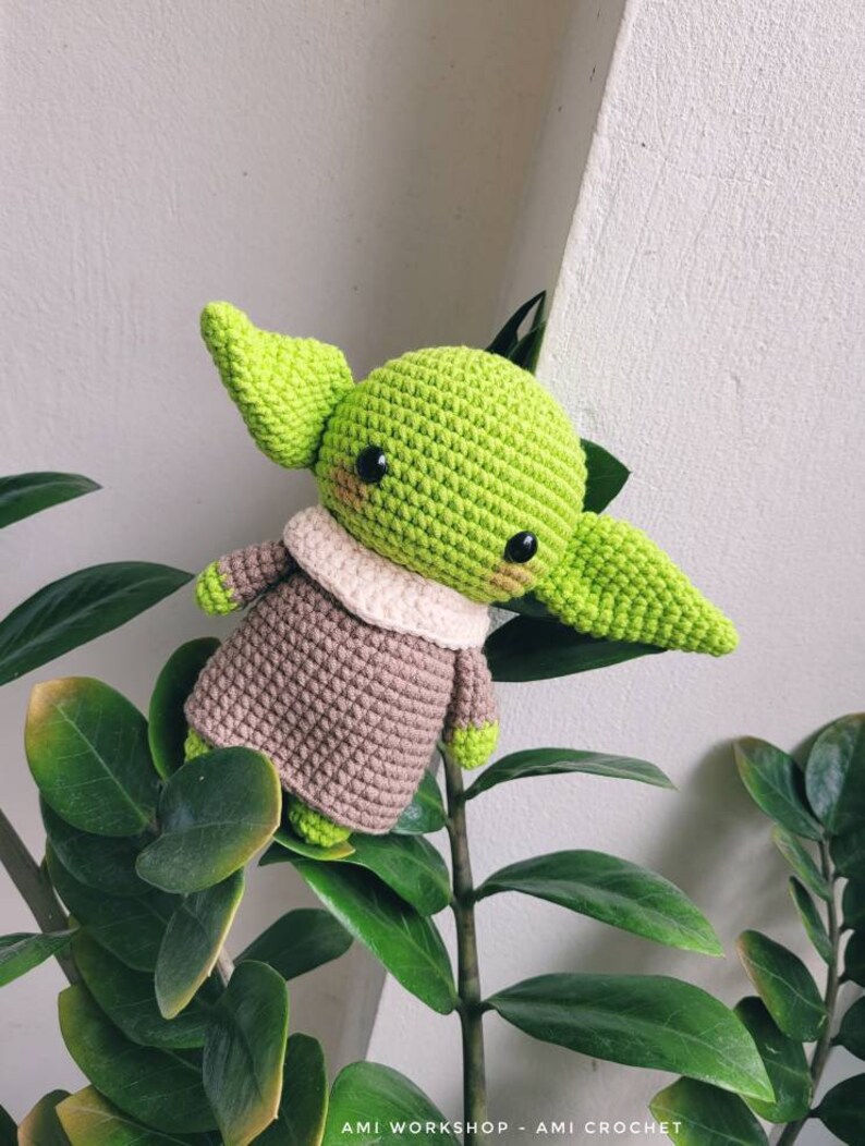 May include: A green crocheted amigurumi toy with brown clothing and large black eyes. The toy is sitting on a green plant.