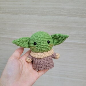 May include: A crocheted green and brown plush toy resembling a character from the Star Wars franchise. The toy has large ears, a small mouth, and black eyes.