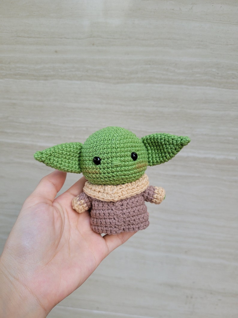 May include: A crocheted green and brown plush toy resembling a character from the Star Wars franchise. The toy has large ears, a small mouth, and black eyes.