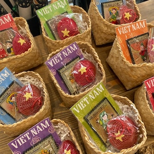 May include: Several small woven baskets filled with gifts. Each basket contains a red crocheted item with a yellow star and a postcard with the text "VIET NAM". The postcards have different colored backgrounds.