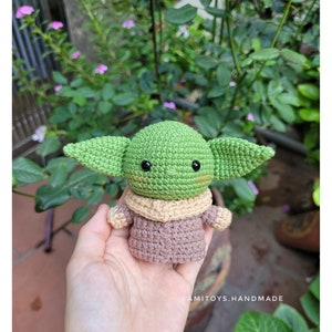 May include: A crocheted green and brown Baby Yoda plush toy with black eyes. The toy is being held in a person's hand.