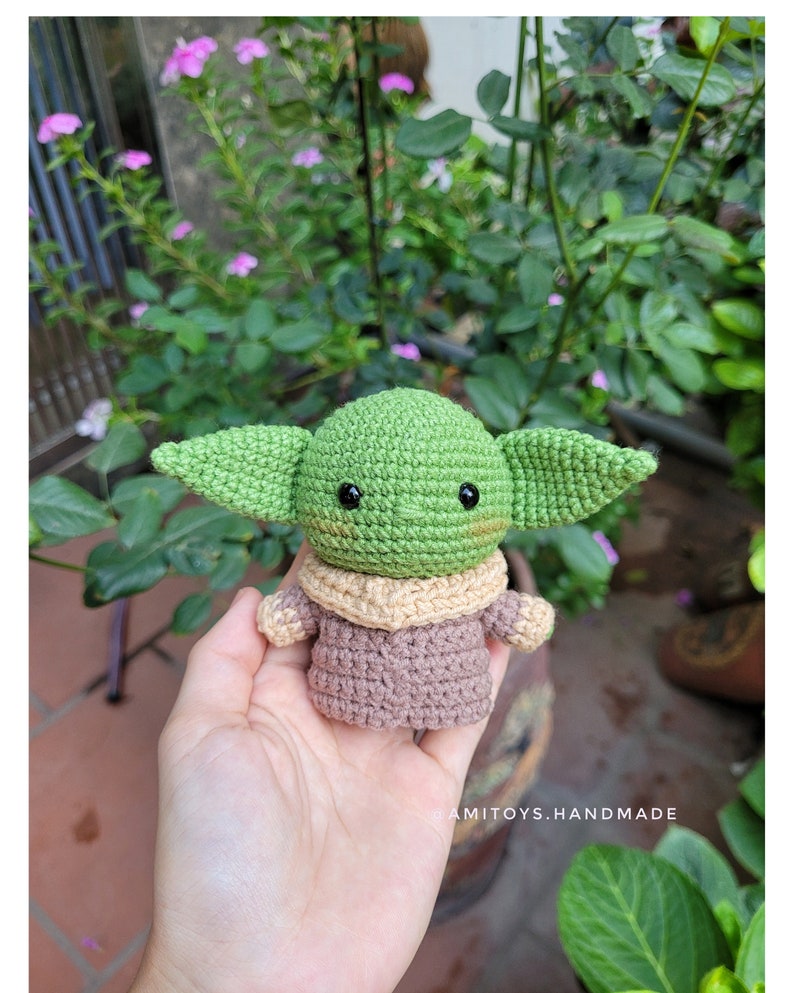 May include: A crocheted green and brown Baby Yoda plush toy with black eyes. The toy is being held in a person's hand.