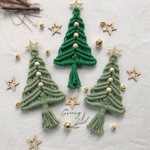 May include: Three green macrame Christmas trees with wooden star tops and white wooden beads. The trees are hanging from a white background with gold bells and stars.