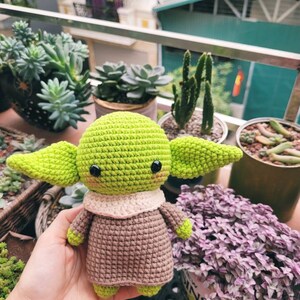 May include: A crocheted green and brown Yoda doll with large eyes and a small mouth. The doll is sitting in a person's hand. The background is a blurry image of plants.