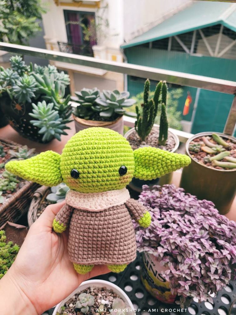 May include: A crocheted green and brown Yoda doll with large eyes and a small mouth. The doll is sitting in a person's hand. The background is a blurry image of plants.