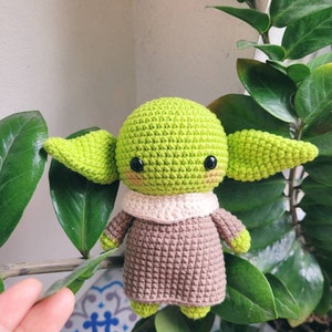 May include: A crocheted green and brown Yoda doll with black eyes and a white collar. The doll is sitting on a green plant.