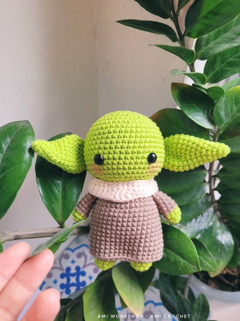 May include: A crocheted green and brown Yoda doll with black eyes and a white collar. The doll is sitting on a green plant.