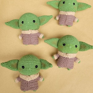 May include: Four crocheted green and brown plush toys shaped like a popular fictional character. The toys have large eyes and small ears. The toys are on a light brown surface. @AMITOYS.HANDMADE