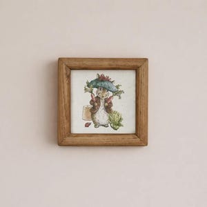 May include: A square wooden frame containing a watercolor illustration of a rabbit wearing a blue hat and brown jacket, holding two carrots. The rabbit stands next to a small terracotta pot, a green leafy vegetable, and a tiny red ladybug.