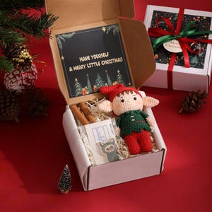 May include: A Christmas gift box with a crocheted elf doll, cinnamon sticks, and a cat magnet. The box has a card that reads "Have yourself a Merry Little Christmas". Another gift box is in the background.