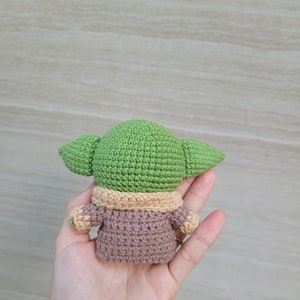 May include: A green and brown crocheted plush toy resembling a character from the Star Wars franchise. The toy is wearing a brown coat with a tan collar.
