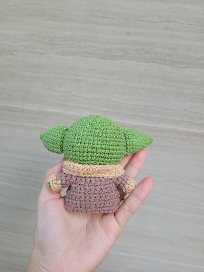 May include: A green and brown crocheted plush toy resembling a character from the Star Wars franchise. The toy is wearing a brown coat with a tan collar.