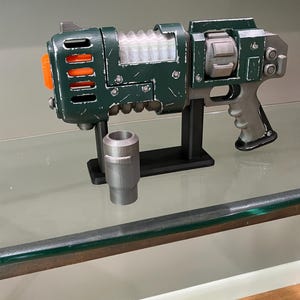 May include: A dark green and grey prop blaster with a weathered finish, featuring a bright orange muzzle tip, a translucent ribbed central section, and a grey handle. The blaster is mounted on a black stand, accompanied by a separate grey cylindrical component.