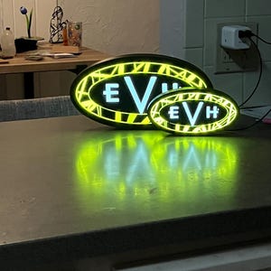 May include: Two illuminated oval signs with the letters "EVH" in white on a black background. The signs have a yellow border with a geometric pattern. The larger sign is in the background and the smaller sign is in the foreground.
