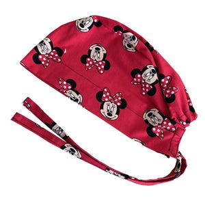 May include: A red scrub cap featuring a repeating pattern of Minnie Mouse's face with a polka dot bow. The cap has two long ties for securing it. The fabric is a vibrant red.