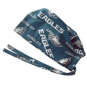 May include: A teal scrub cap featuring the word "EAGLES" in white block letters and the team logo. The cap has a tie closure and is made of a patterned fabric. The cap is designed for medical professionals or sports fans.