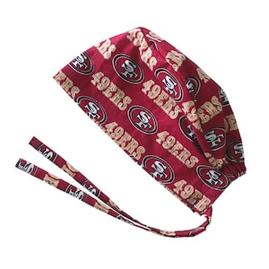 May include: A maroon scrub cap featuring a repeating pattern of the San Francisco 49ers logo and the text 49ERS in gold and white. The cap includes two long fabric ties at the back for securing the fit, with a gathered design at the nape of the neck.