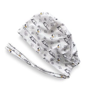 May include: A white scrub cap featuring a repeating pattern of Snoopy characters and grey paw prints. The cap includes long fabric ties for securing the fit and has a gathered back section to accommodate various hair lengths and styles comfortably.