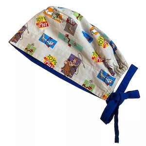 May include: A white surgical scrub cap with a blue lining and ties. The cap is patterned with cartoon characters and the words "TOYS at PLAY" in red and yellow.