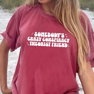 May include: A maroon oversized t-shirt with the text "Somebody's Crazy Conspiracy Theorist Friend" printed in white.