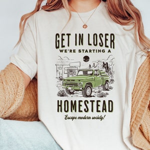 May include: White t-shirt with a green truck graphic and the text "Get in loser we're starting a homestead Escape modern society!"