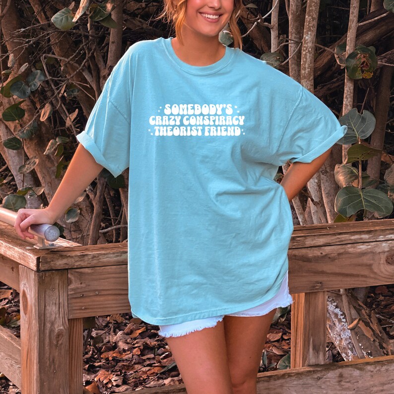 May include: A light blue t-shirt with the text "Somebody's Crazy Conspiracy Theorist Friend" in white, retro-style lettering.