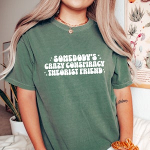 May include: A green t-shirt with the text "Somebody's Crazy Conspiracy Theorist Friend" in white, retro-style lettering.