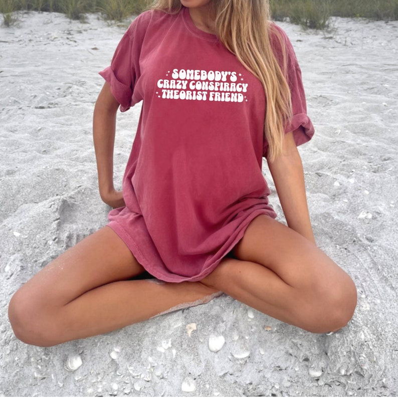 May include: A maroon oversized t-shirt with the text "Somebody's Crazy Conspiracy Theorist Friend" printed in white.