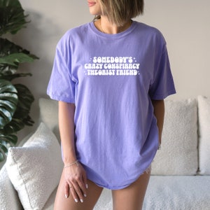 May include: A purple t-shirt with the text "Somebody's Crazy Conspiracy Theorist Friend" in white letters.
