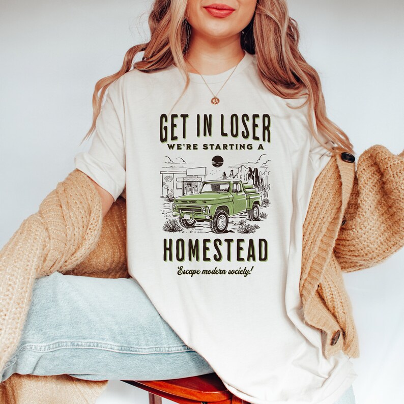 May include: White t-shirt with a green truck graphic and the text "Get in loser we're starting a homestead Escape modern society!"