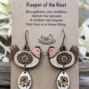 May include: Wooden earrings featuring a hen design with a floral pattern and an egg-shaped dangle. The earrings are attached to a card with the text "Keeper of the Nest" and a poem about a mother hen.