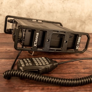 Pode incluir: Um transceptor de rádio preto com um microfone e um cabo enrolado. O rádio é etiquetado como "IC-7000" e está montado em uma gaiola de metal preta etiquetada como "XTenna".