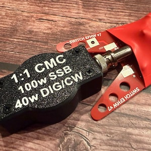May include: A black 1:1 CMC device with the text "100w SSB 40w DIG/CW" and a silver connector. A red circuit board labeled "SWITCH EFHW V7" is partially visible. The device is on a wooden surface.