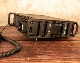 May include: A black radio transceiver with a large knob and a digital display. The radio is mounted in a black protective case with the text "FT 710" and "xTenna" printed on it. The radio is sitting on a wooden surface.