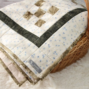 Peut inclure: Une courtepointe en patchwork blanc et brun avec un motif floral et une bordure noire et verte. La courtepointe est pliée et placée dans un panier en osier.