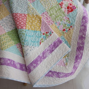 Könnte beinhalten: Eine Patchwork-Decke mit weißem Hintergrund und verschiedenen bunten Blumenmustern. Die Decke ist mit einem hellblauen Saum und einem violetten Band eingefasst.