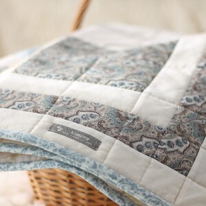Peut inclure: Une courtepointe blanche et bleue en patchwork avec un motif paisley. La courtepointe est pliée et placée dans un panier en osier. La courtepointe porte une étiquette indiquant "Kimera".
